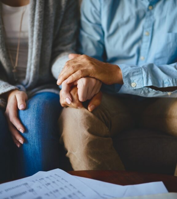 photo of a man and woman seated in front a several pieces of paper while holding hands