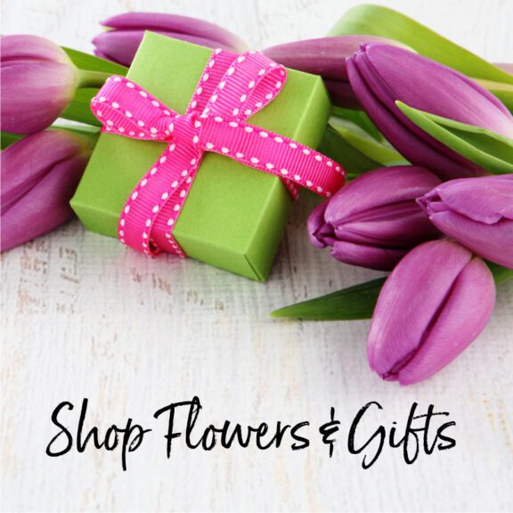 Photo of a small wrapped box with purple flowers surrounding it and the words Shop Flowers-Gifts underneath