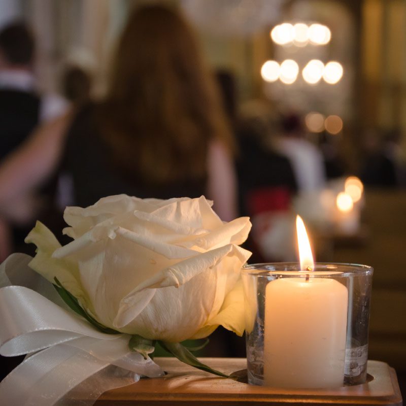 Closeup of lit candle and memorial flower in the forefront of Funeral Celebrant speaking to loved ones