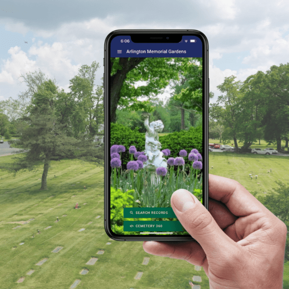 A person's hand holding up a phone showing the Locate a Loved One mobile app with a view of a cemetery in the background
