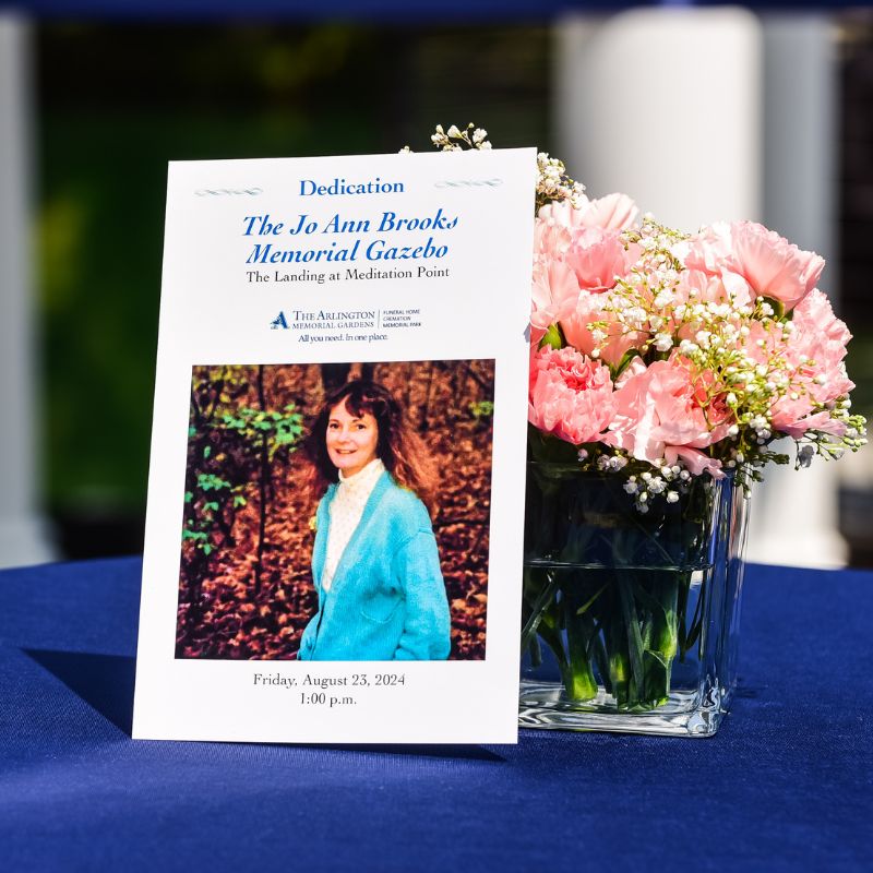 Brooks Dedication Program with a photo of Jo Ann Brooks on it leaning against a small vase of pink and white flowers