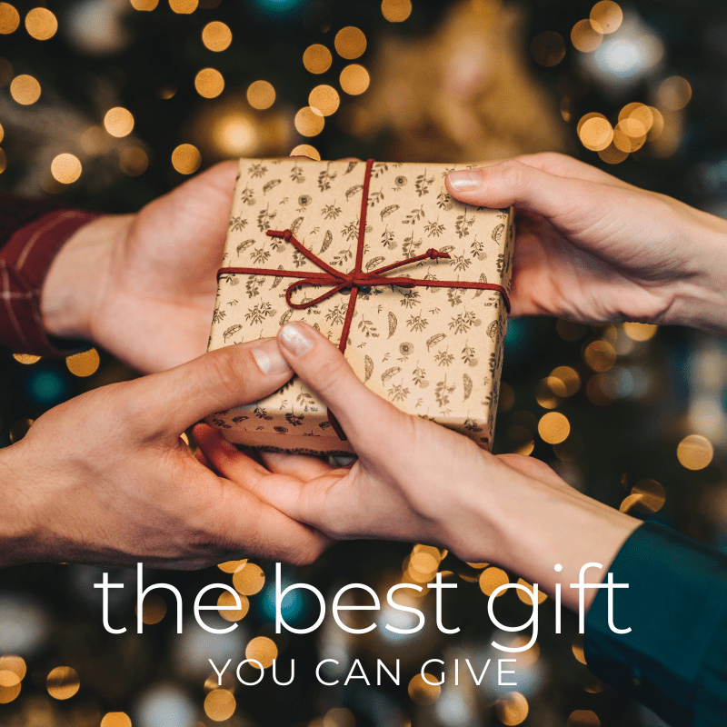 Photo of two people passing a small wrapped Christmas present in their hands with a Christmas tree in the background and text that says "the best gift you can give"